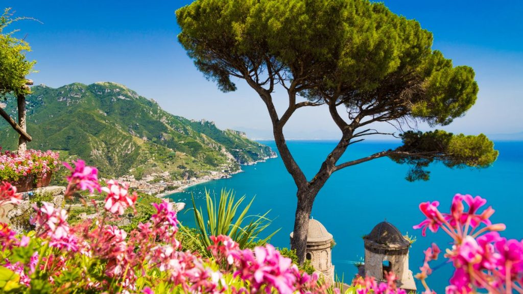 Panormablick über die grüne Landschaft, das kristallblaue Meer und die kleinen Häuser der italienischen Amalfiküste. Umrandet von roten und pinken Blumen